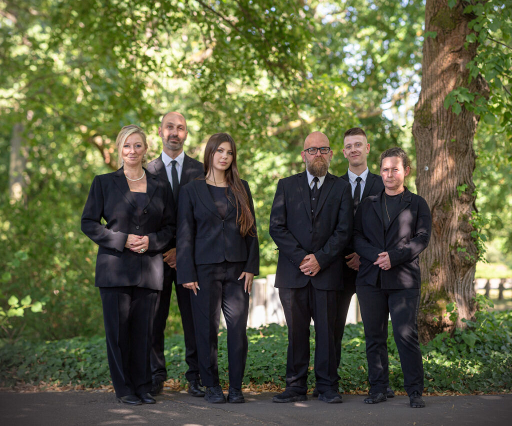 Albatros-Bestattungen® Team steht aufgereiht mit schwarzer Kleidung vor Bäumen